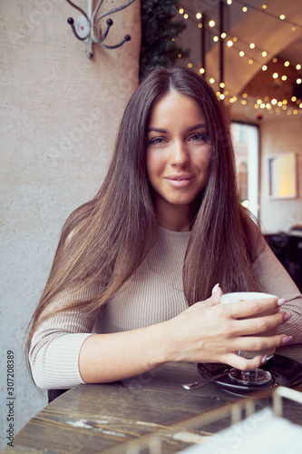 Beautiful pensive happy girl sitting in cafe in Christmas holidays, smiling and dreaming. Brunette woman with long hair drinks cappuccino coffee, latte and looks to camera