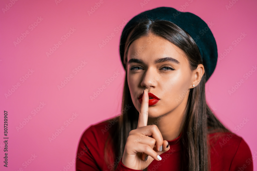 Beautiful woman with red lips holding finger on her lips over pink background. Gesture of shhh, secret, silence. Close up. Beautiful woman with red lips holding finger on her lips over pink background. Gesture of shhh, secret, silence. Close up.
