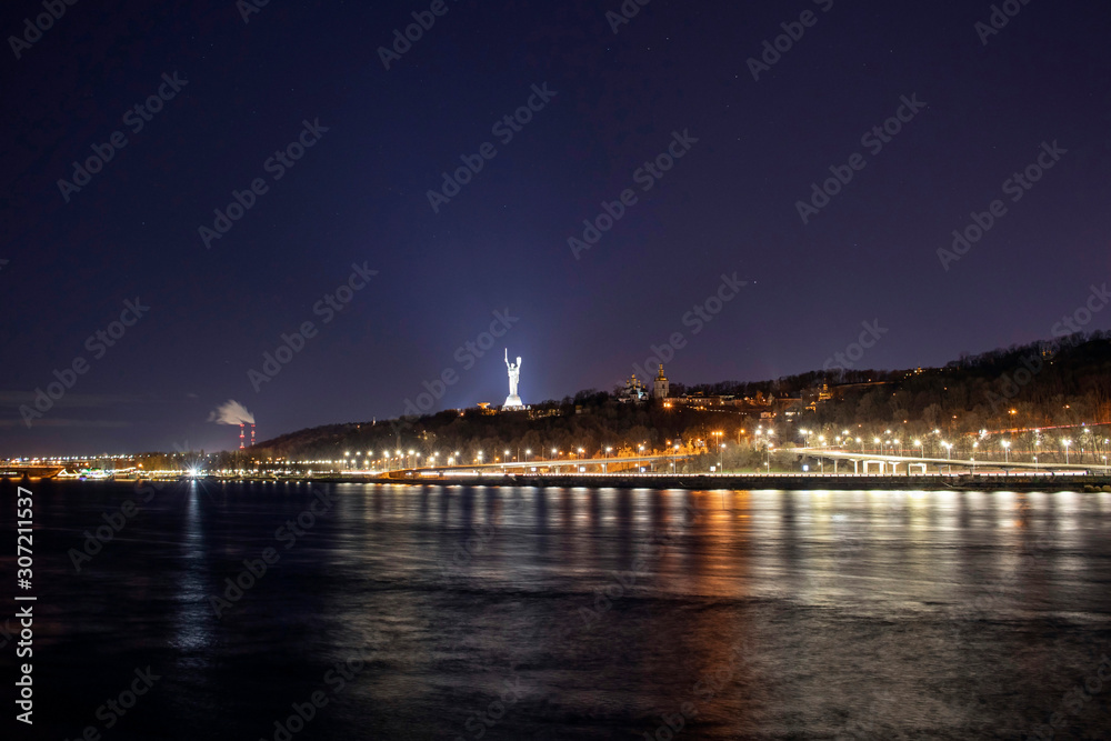 Beautiful night city of Kiev and the Dnieper river in the foreground with illumination of the embankment with a long exposure, Kiev, Ukraine