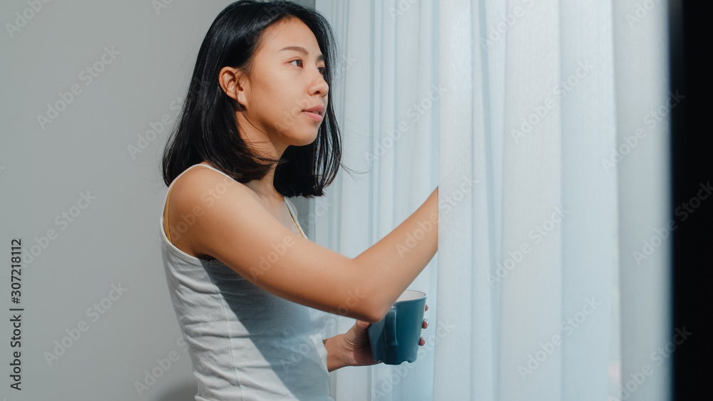 Happy beautiful Asian woman smiling and drinking a cup of coffee or tea near the window in bedroom. Young latin girl open curtains and relax in morning. Lifestyle lady at home concept. Happy beautiful Asian woman smiling and drinking a cup of coffee or tea near the window in bedroom. Young latin girl open curtains and relax in morning. Lifestyle lady at home concept.