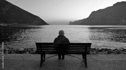 A lonely old man sits on a wooden bench at dusk after sunset, looking at the lake and the light on the horizon.   photo