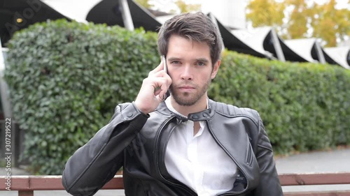 Attractive young man looks seriously at camera while talking through his smartphone. Outdoor