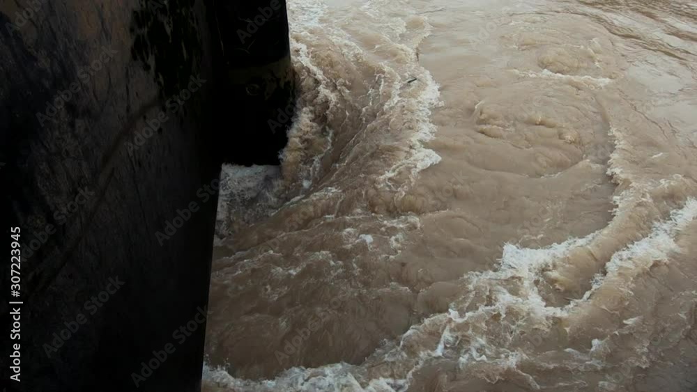Slow motion flooded river filled with brown dirty water spilling under floodgate Slow motion flooded river filled with brown dirty water spilling under floodgate
