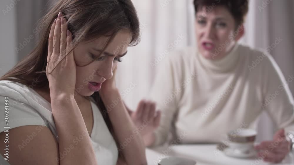 Side view of adult Caucasian pretty girl holding hands on ears as senior woman yelling at the background. Family relationship, problem, confrontation, ignoring. Side view of adult Caucasian pretty girl holding hands on ears as senior woman yelling at the background. Family relationship, problem, confrontation, ignoring.
