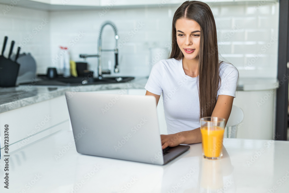 Young smiling woman having breakfast in the kitchen connecting with a laptop and drinking an healthy orange juice Young smiling woman having breakfast in the kitchen connecting with a laptop and drinking an healthy orange juice