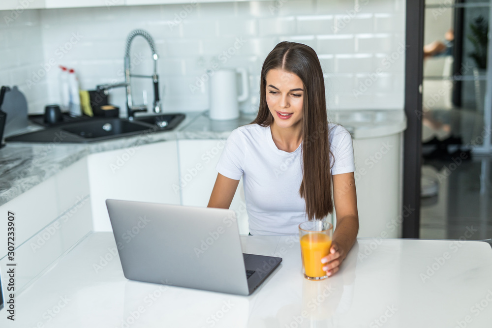 Young smiling woman having breakfast in the kitchen connecting with a laptop and drinking an healthy orange juice Young smiling woman having breakfast in the kitchen connecting with a laptop and drinking an healthy orange juice