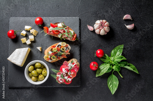 Traditional Bruschetta. Italian antipasti set with jamon, guanchial sausage, olives, cottage cheese, arugula and tomatoes on a black background