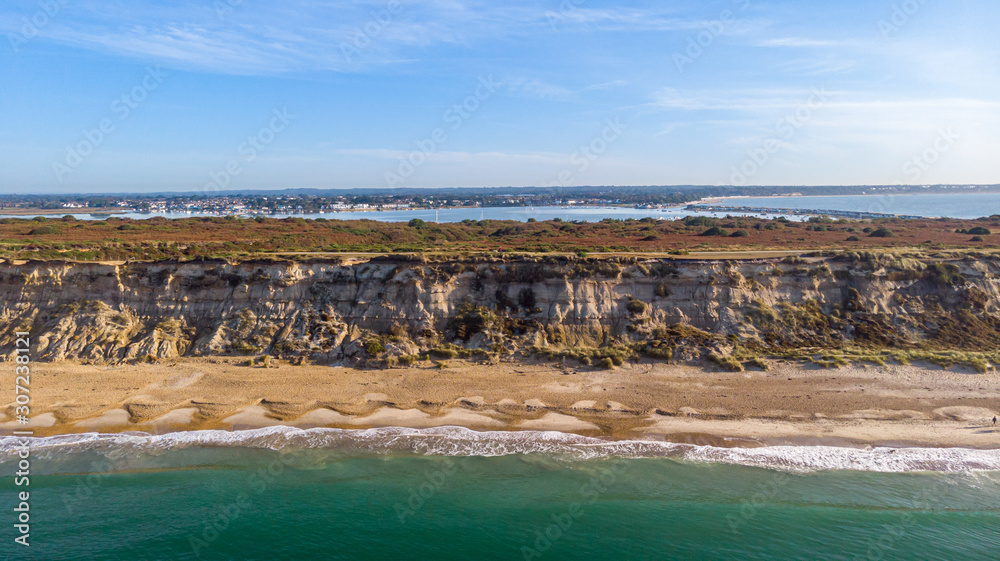 An aerial view of a majestic seacoast hill with huge cliff, sandy beach, groyne (breakwater) and crystal blue water under a beautiful blue sky and some white clouds