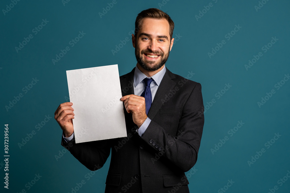 Businessman holding white vertical a4 paper poster. Copy space. Smiling handsome man in corporate suit with tie on blue background. Businessman holding white vertical a4 paper poster. Copy space. Smiling handsome man in corporate suit with tie on blue background.