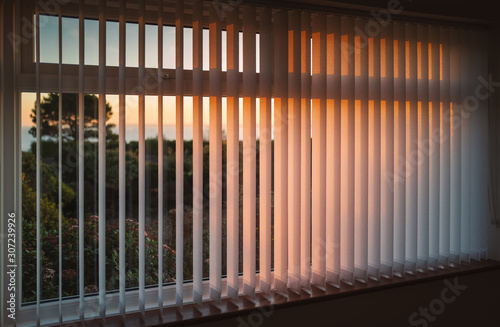White vertical slat blinds hanging in front of a window as the sun is setting turning the light golden. The slats have sealed glued pockets and no cords at the bottom.