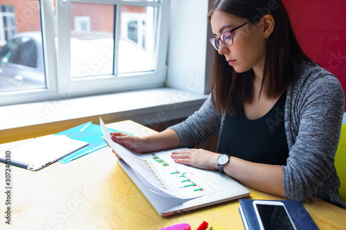 Serious woman in glasses successful economist reading paper documents with financial schedules while sitting at wooden table in office interior. Female business worker reading graph