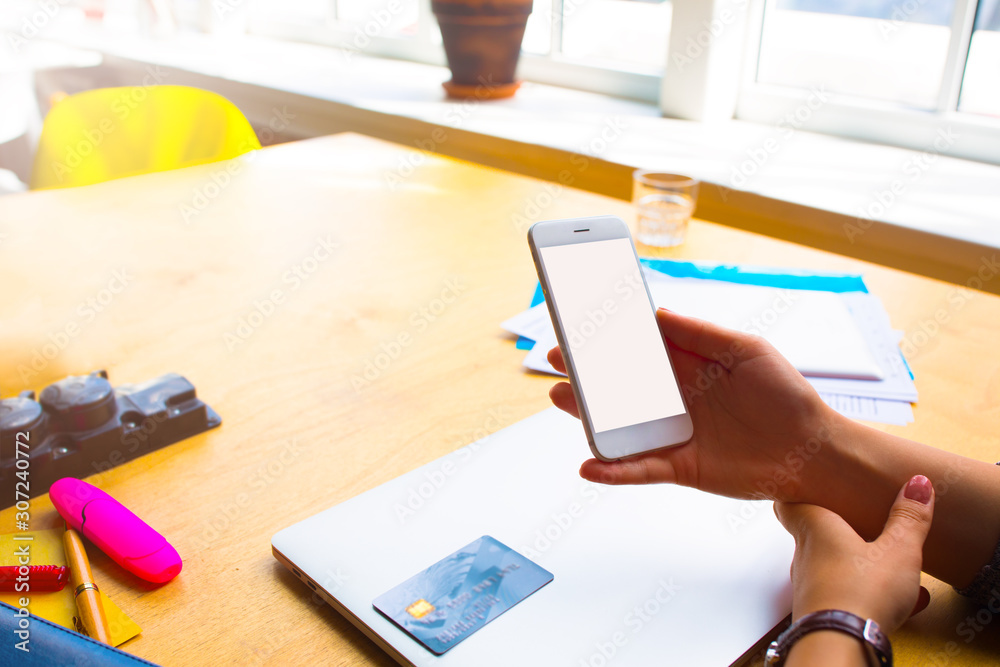 Woman financier online booking via cellphone with blank screen and using for payment credit debit card while sitting at desktop with closed netbook in modern office interior