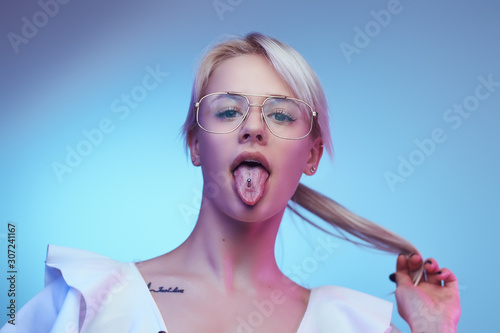 Closeup portrait of a cocky blonde girl wearing glasses poses with tongue sticking out looking at the camera. Isolated on a blue background