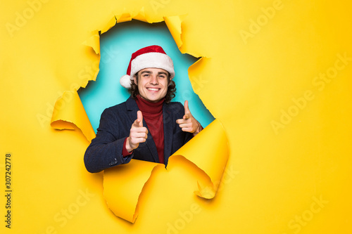 Young handsome man in santa hat pointed with hands you on copy space through yellow paper hole. Christmas sales. © F8  \ Suport Ukraine