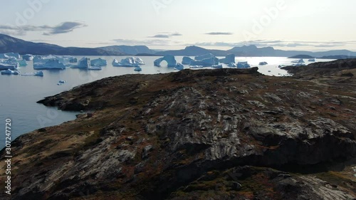 Slow drone fly-by view of Akukkeq island, Greenland, flying over a highly detailed dark rocky terrain from this virgin and desertic island, revealing floating Icebergs in the background