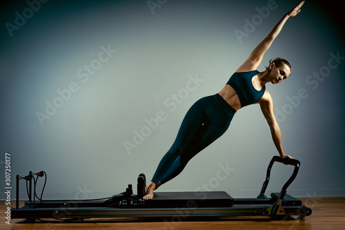Young girl doing pilates exercises with a reformer bed. Beautiful slim fitness trainer on reformer gray background, low key, art light. Fitness concept