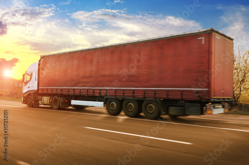 A big white truck and trailer driving fast on a countryside road against a sky with a sunset