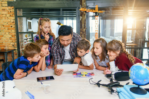 Foto Group of curious school children listen their Korean male teacher uses device of tablet computer for show educational films for pupils