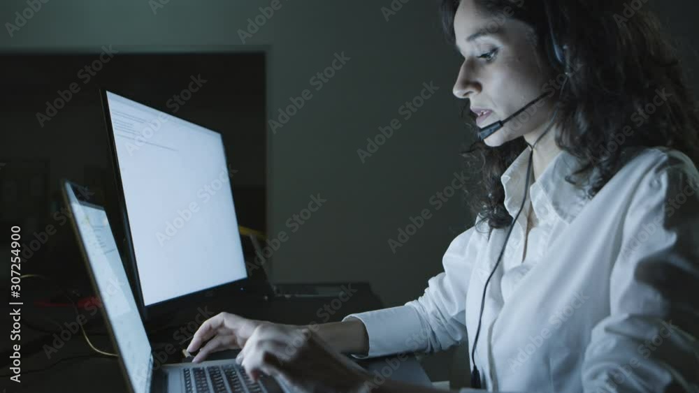 Teleworker talking and typing on laptop. Side view of young female call center operator in headset using computer at night. Customer service concept