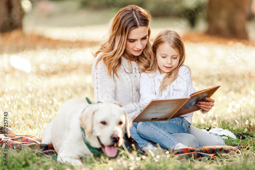 Happy beautiful mother and daughter with dog labrador is reads book sitting in the park. © zadorozhna