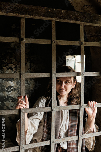 A young woman in a vintage dress from the 18-19 century looks through the bars of a prison cell.