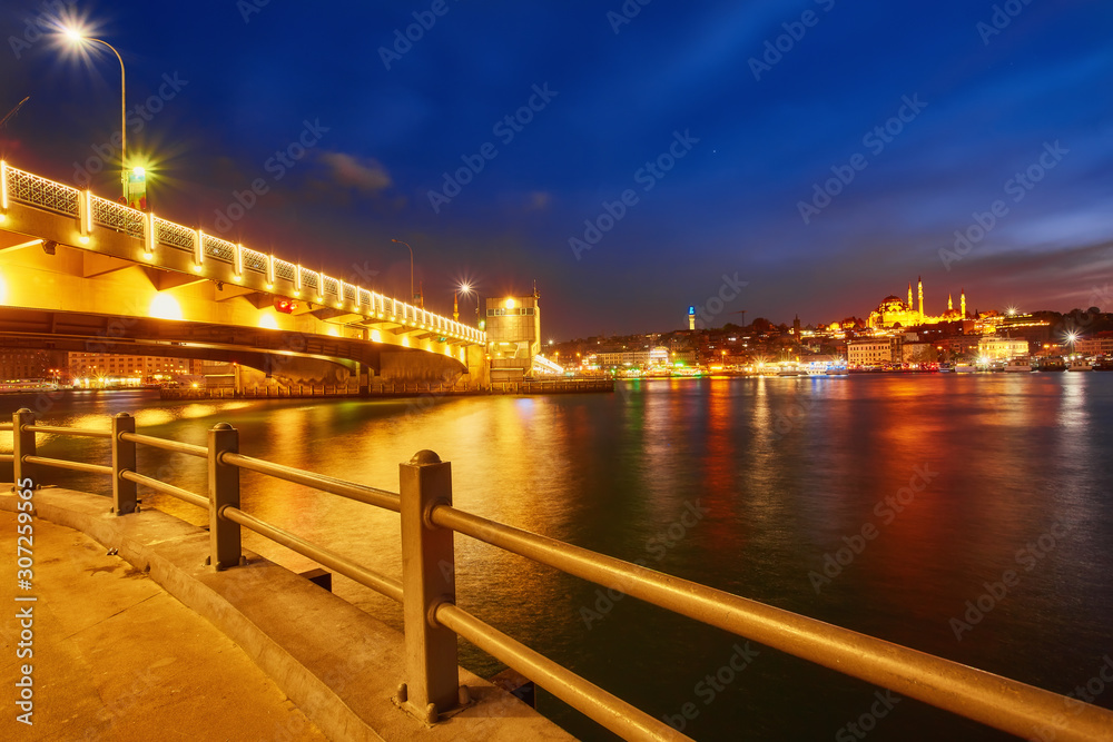 Fototapeta premium Night view at Yeni Cami Mosque worship place from Galata bridge reflected in water of Golden Horn of Bosporus.