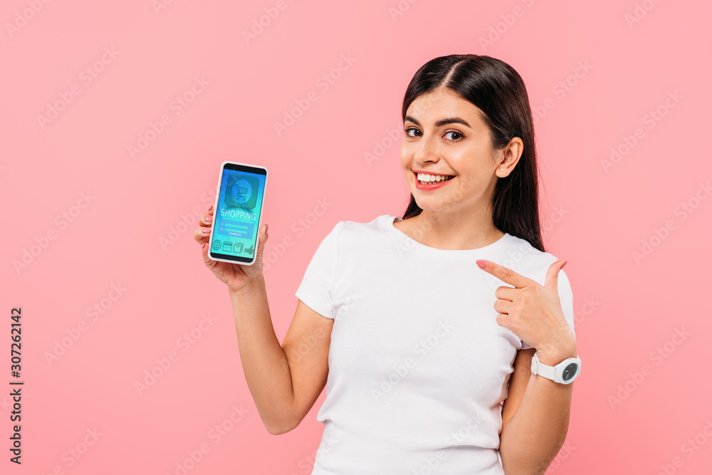smiling pretty brunette girl pointing with finger at smartphone with online shopping app isolated on pink smiling pretty brunette girl pointing with finger at smartphone with online shopping app isolated on pink