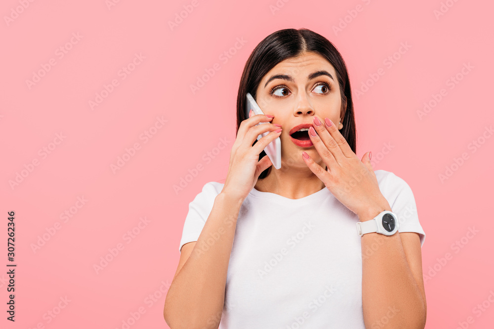 shocked pretty brunette girl talking on smartphone isolated on pink