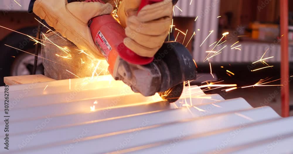 A construction worker using a power tool angle grinder to cut metal and send glowing bright sparks flying in slow motion.