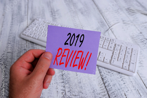 Text sign showing 2019 Review. Business photo showcasing remembering past year events main actions or good shows man holding colorful reminder square shaped paper white keyboard wood floor