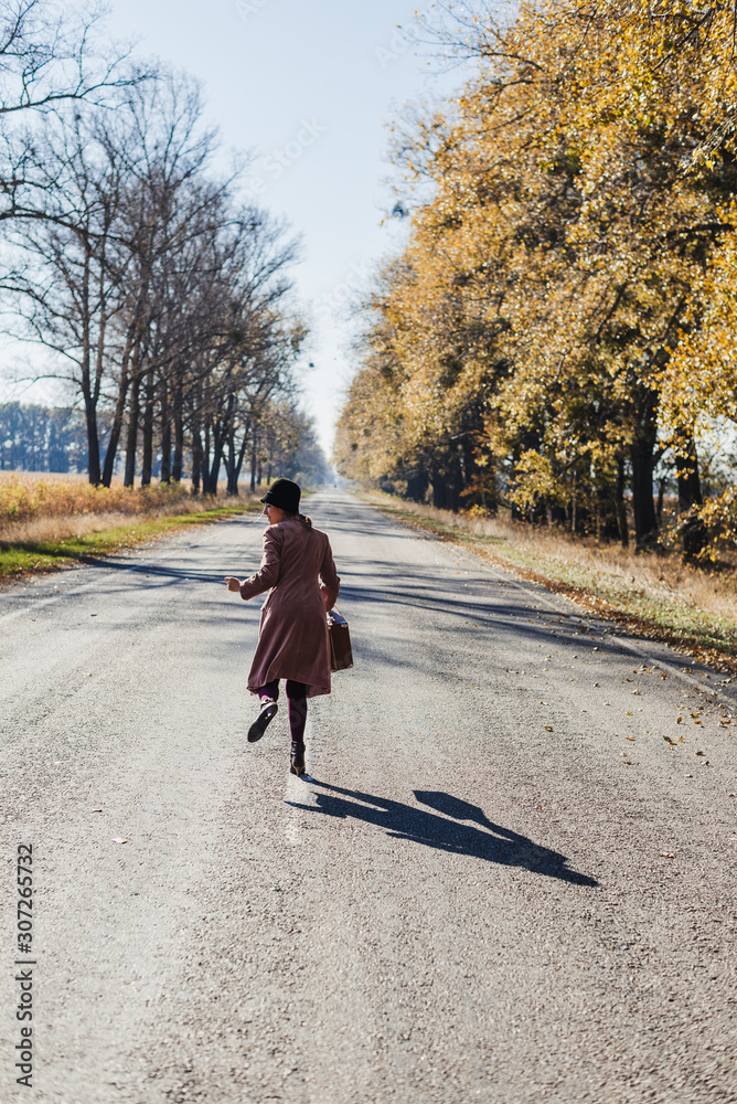 Young redhead lady woman in pink vintage coat and hat with suitcase in retro style running away along a park road with golden yellow autumnal trees. Outdoor autumn garden relaxation, travel concept