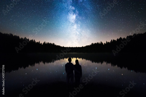 Fotografie, Obraz Romantic Couple Standing Under The Starry Sky, Milky Way Reflects Off Lake