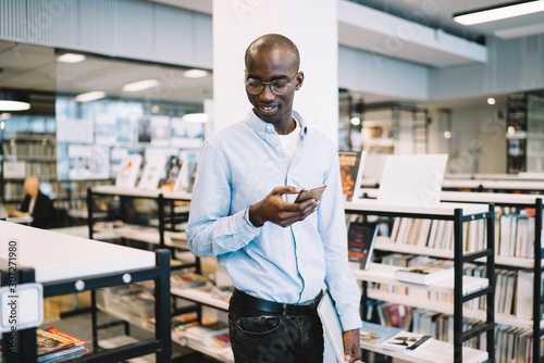 Energy black man walking fast along bookcases with smartphone