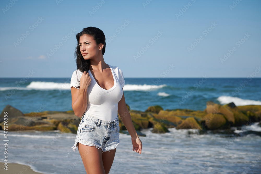 Stunning young brunette woman wearing white t-shirt and denim shorts on beach Stunning young brunette woman wearing white t-shirt and denim shorts on beach