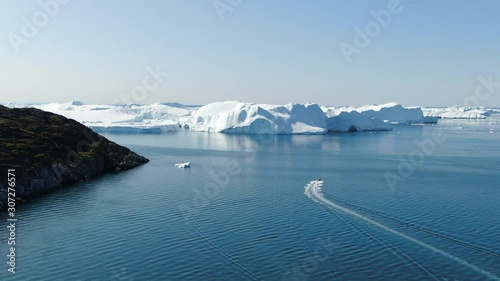 Slow drone aerial view of the area of Ilulissat, Greenland, following a small boat cruising in the calm, blue Baffin Sea, facing majestic ice bank and small icebergs in the background