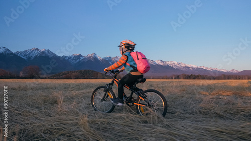 One caucasian children rides bike in wheat field. Little girl riding black orange cycle on background of beautiful snowy mountains. Biker motion ride with backpack and helmet. Mountain bike hardtail.