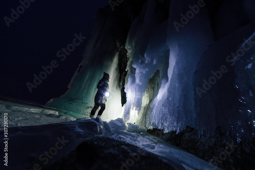 Surreal landscape with woman exploring mysterious ice grotto cave. Outdoor adventure. Girl exploring huge icy cave, dark majestic landscape. Magical silhouettes on background of illuminated ice blocks