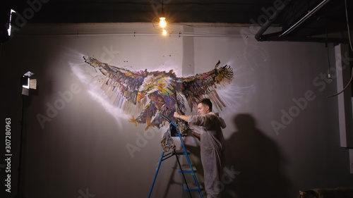 Artist designer draws an eagle on the wall. Craftsman decorator paints a picture with acrylic oil color. Painter painter dressed in a paint coat. Indoor. Dark magic cinematic look. photo