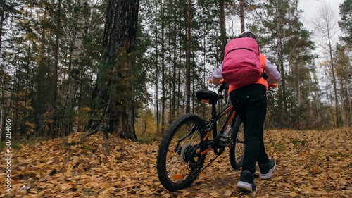 One caucasian children walk with bike in autumn park. Little girl walking black orange cycle in forest. Kid goes do bicycle sports. Biker motion ride with backpack and helmet. Mountain bike hardtail.