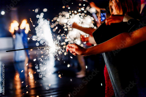 A crowd of young happy people with sparklers in their hands during celebration. Sparkler in hands on a wedding 2020 - bride, groom and guests holding lights in hand. Sparkling lights of bengal fires.