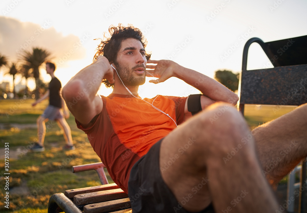 Confident young man listening to music in headphones from smart phone armband doing exercises on public equipment in the outdoor gym at the park - obrazy, fototapety, plakaty 