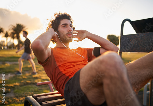 Fototapeta Naklejka Na Ścianę i Meble -  Confident young man listening to music in headphones from smart phone armband doing exercises on public equipment in the outdoor gym at the park