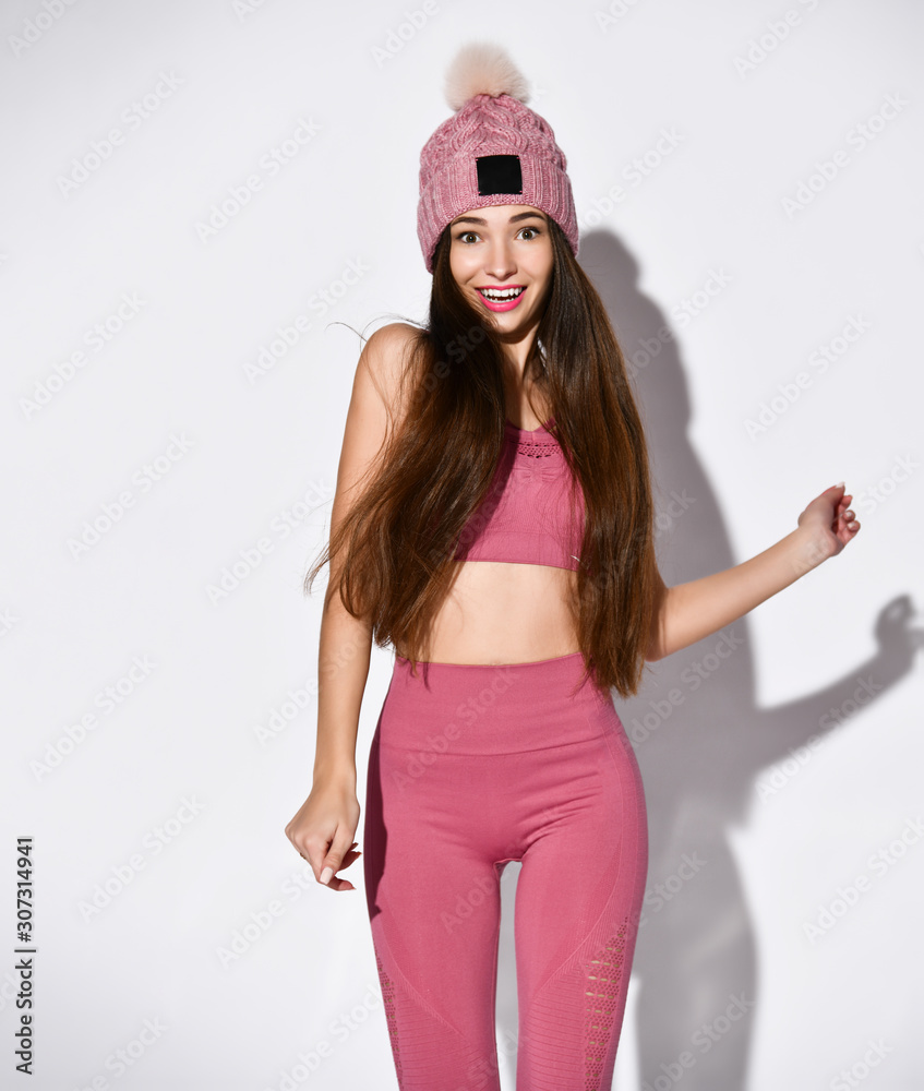 Foto de Athletic young woman in sports attire, tight-fitting pink leggings  and top s color, warm knitted hat with fur pompom, in motion, fitness,  isolated in studio do Stock