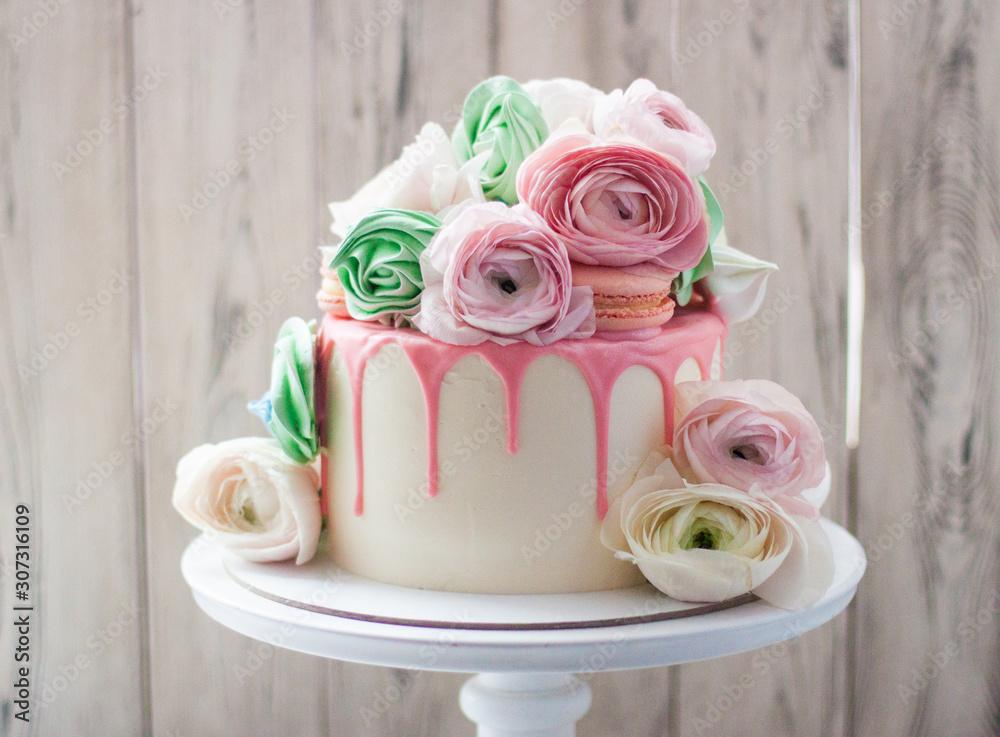 White cake with pink melted chocolate decorated with fresh flowers, roses, ranunculuses, macaroons and meringue swirls. 