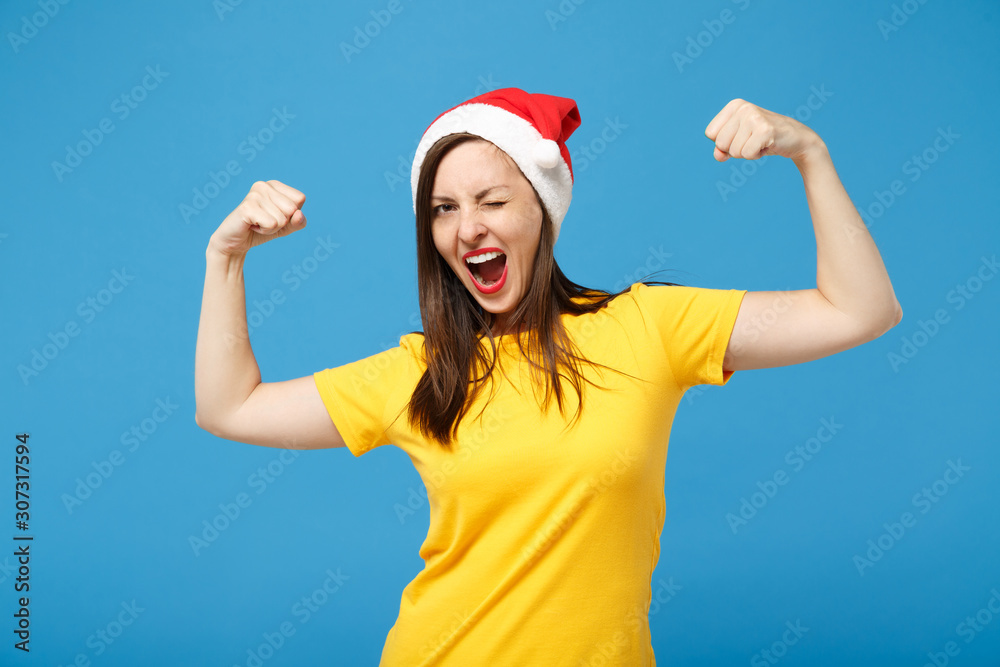 Funny young woman Santa girl in yellow t-shirt, Christmas hat posing isolated on blue background. Happy New Year 2020 celebration holiday concept. Mock up copy space. Showing biceps, muscles blinking. Funny young woman Santa girl in yellow t-shirt, Christmas hat posing isolated on blue background. Happy New Year 2020 celebration holiday concept. Mock up copy space. Showing biceps, muscles blinking.