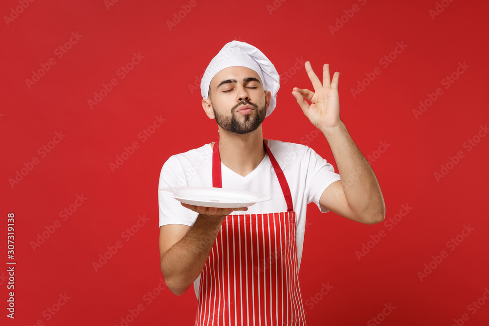 Chef cook or baker man in striped apron toque chefs hat isolated on red background. Cooking food concept. Mock up copy space. Hold empty blank plate with place for food making okay taste delight sign. Chef cook or baker man in striped apron toque chefs hat isolated on red background. Cooking food concept. Mock up copy space. Hold empty blank plate with place for food making okay taste delight sign.