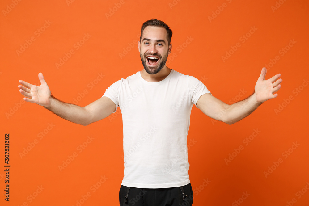 Excited surprised young man in casual white t-shirt posing isolated on bright orange wall background studio portrait. People sincere emotions lifestyle concept. Mock up copy space. Spreading hands. Excited surprised young man in casual white t-shirt posing isolated on bright orange wall background studio portrait. People sincere emotions lifestyle concept. Mock up copy space. Spreading hands.