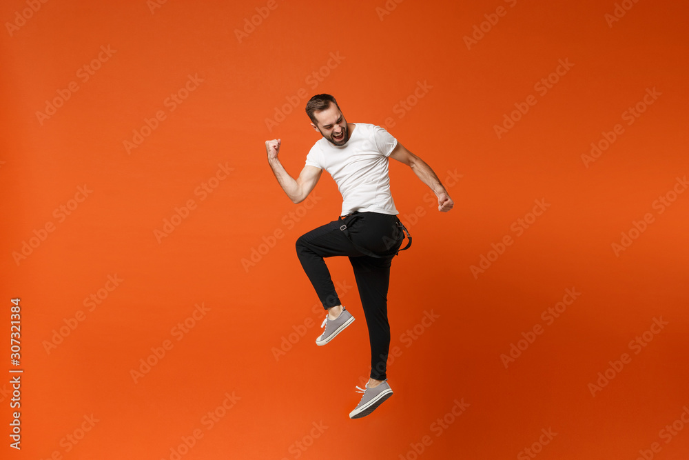 Cheerful young man in casual white t-shirt posing isolated on bright orange wall background studio portrait. People lifestyle concept. Mock up copy space. Having fun, jumping, doing winner gesture.