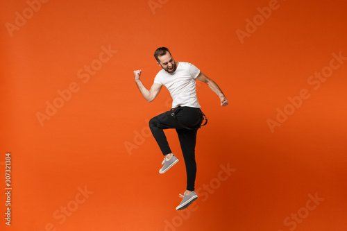 Cheerful young man in casual white t-shirt posing isolated on bright orange wall background studio portrait. People lifestyle concept. Mock up copy space. Having fun, jumping, doing winner gesture.