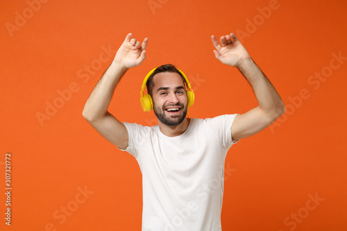 Handsome funny young man in casual white t-shirt posing isolated on orange wall background studio portrait. People lifestyle concept. Mock up copy space. Listening music with headphones, dancing.